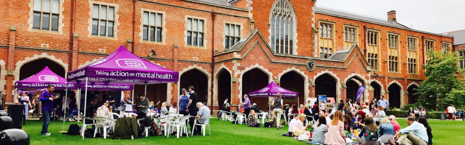 Image shows Action Mental Health Picnic taking place on the front lawn of Queen's. Staff are having lunch on white plastic chairs on a sunny day.